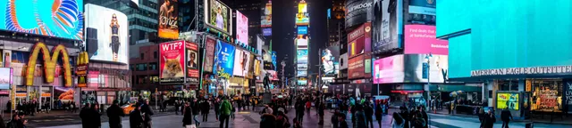 Panorama of a busy city square surrounded by illuminated billboards at night.