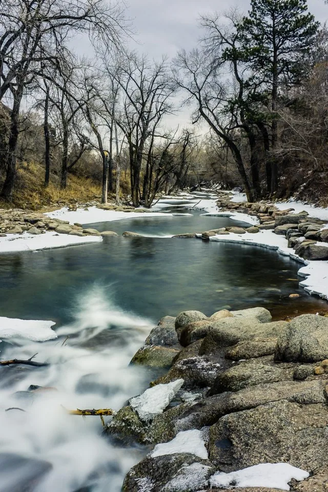 A stream flows over rocks through snow-covered woodland.
