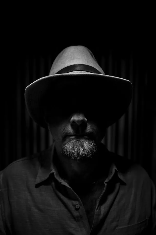 Black-and-white portrait of a bearded person wearing a brimmed hat in deep shadow.