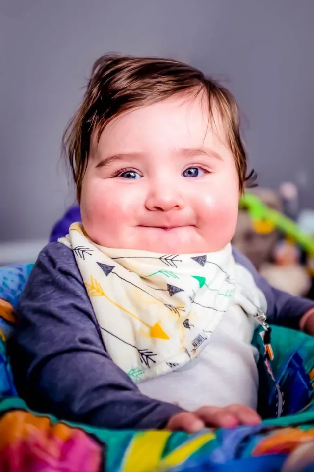 A smiling baby wearing a patterned bib sits among colorful blankets.