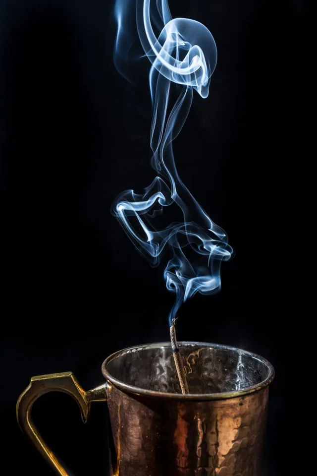 Blue-white smoke rises from a metal mug against black.
