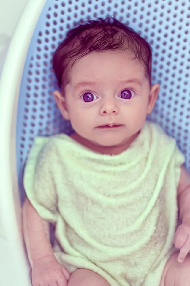 A wide-eyed baby in a pale green bib lies on a patterned blanket.