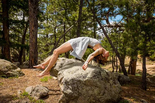 A person arches backward above a large boulder in a sunlit pine forest.